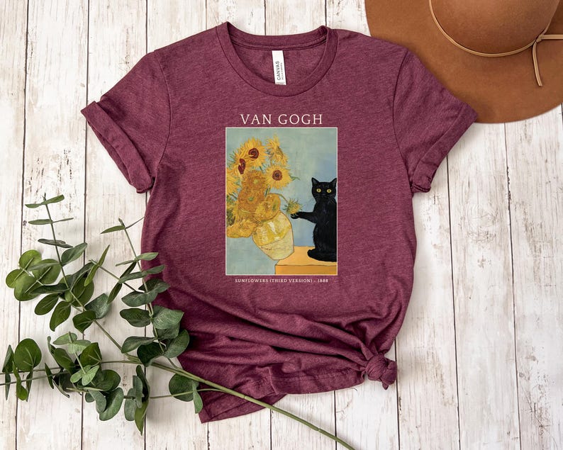 May include: Burgundy t-shirt featuring a print of Van Gogh's "Sunflowers" painting with a black cat. The text "VAN GOGH" is above the image, and "SUNFLOWERS (THIRD VERSION) 1888" is below. The shirt has short sleeves and a crew neck.