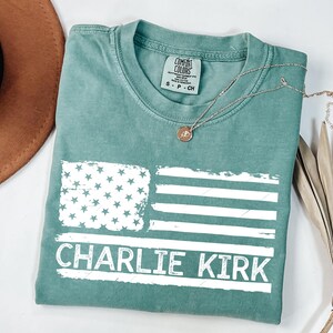 Peut inclure: Un t-shirt Comfort Colors de couleur sarcelle avec un drapeau am&eacute;ricain blanc vieilli et le nom "Charlie Kirk" imprim&eacute; en dessous. Un collier argent&eacute; avec un pendentif est drap&eacute; sur le t-shirt. Le t-shirt est pli&eacute; sur une surface blanche.