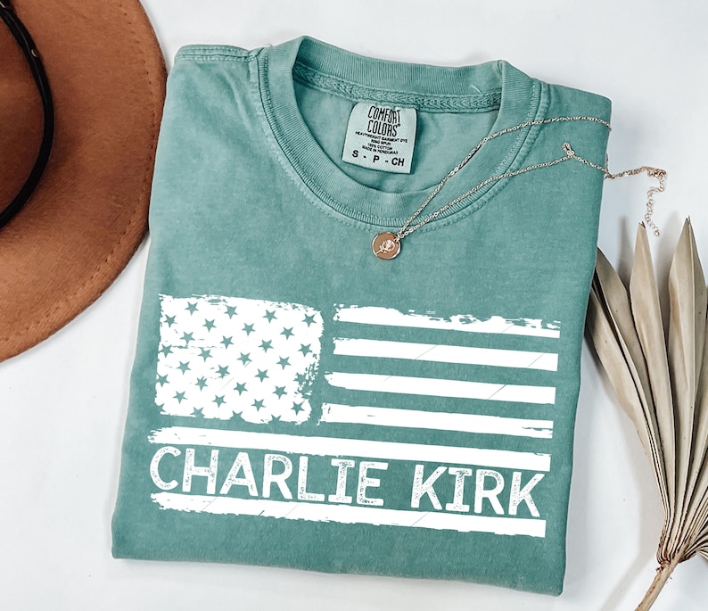 Peut inclure: Un t-shirt Comfort Colors de couleur sarcelle avec un drapeau am&eacute;ricain blanc vieilli et le nom "Charlie Kirk" imprim&eacute; en dessous. Un collier argent&eacute; avec un pendentif est drap&eacute; sur le t-shirt. Le t-shirt est pli&eacute; sur une surface blanche.