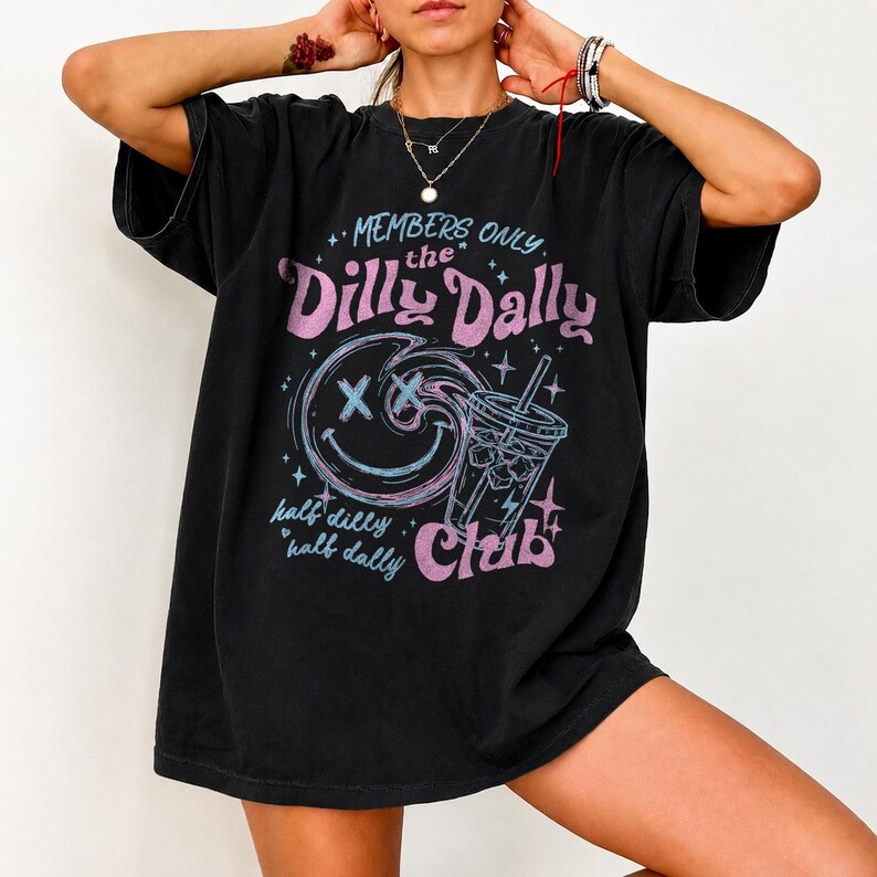 May include: A black short-sleeve t-shirt featuring a graphic print with the text Members Only the Dilly Dally Club and half dilly half dally. The design includes a blue swirling smiley face and a blue iced beverage cup with pink accents.