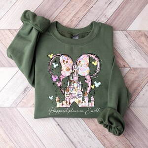 K&ouml;nnte beinhalten: Ein olivgr&uuml;nes Sweatshirt mit einem farbenfrohen Aufdruck eines Schlosses und Mickey Mouse Ohren. Das Design beinhaltet Pastellfarben und den Text "Happiest place on Earth". Das Sweatshirt hat lange &Auml;rmel mit gerippten B&uuml;ndchen.