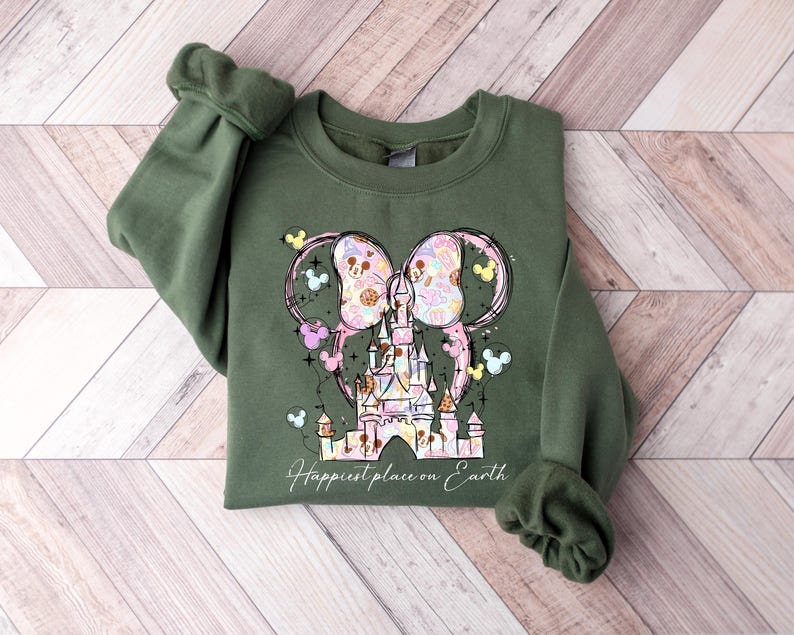 K&ouml;nnte beinhalten: Ein olivgr&uuml;nes Sweatshirt mit einem farbenfrohen Aufdruck eines Schlosses und Mickey Mouse Ohren. Das Design beinhaltet Pastellfarben und den Text "Happiest place on Earth". Das Sweatshirt hat lange &Auml;rmel mit gerippten B&uuml;ndchen.