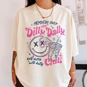 May include: A cream-colored short-sleeve t-shirt featuring a graphic print with the text "MEMBERS ONLY the Dilly Dally Club" and "half dilly half dally". The design includes a pink and blue smiley face with X eyes and a drink cup with ice.