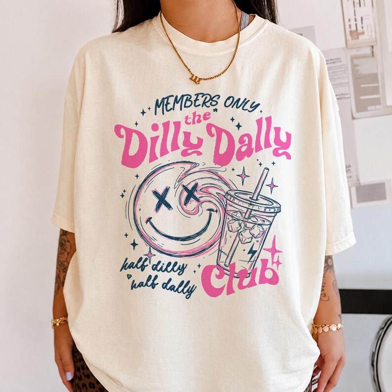May include: A cream-colored short-sleeve t-shirt featuring a graphic print with the text "MEMBERS ONLY the Dilly Dally Club" and "half dilly half dally". The design includes a pink and blue smiley face with X eyes and a drink cup with ice.