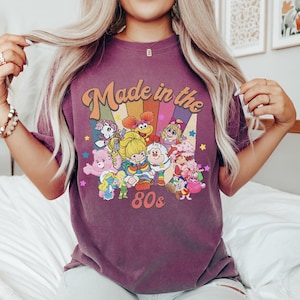 Pode incluir: Uma t-shirt de manga curta roxa com uma impress&atilde;o gr&aacute;fica com o texto Made in the 80s. O design inclui um fundo de raios de sol coloridos e v&aacute;rias personagens de desenhos animados, incluindo um Ursinho Carinhoso, Rainbow Brite e um Smurf.