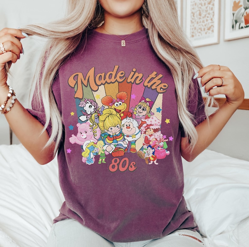 Pode incluir: Uma t-shirt de manga curta roxa com uma impress&atilde;o gr&aacute;fica com o texto Made in the 80s. O design inclui um fundo de raios de sol coloridos e v&aacute;rias personagens de desenhos animados, incluindo um Ursinho Carinhoso, Rainbow Brite e um Smurf.