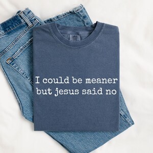 Pu&ograve; includere: Una t-shirt girocollo color blu ardesia con la scritta bianca "I could be meaner but jesus said no" stampata sul davanti, abbinata a un paio di jeans blu. La maglietta ha maniche corte e una vestibilit&agrave; standard.