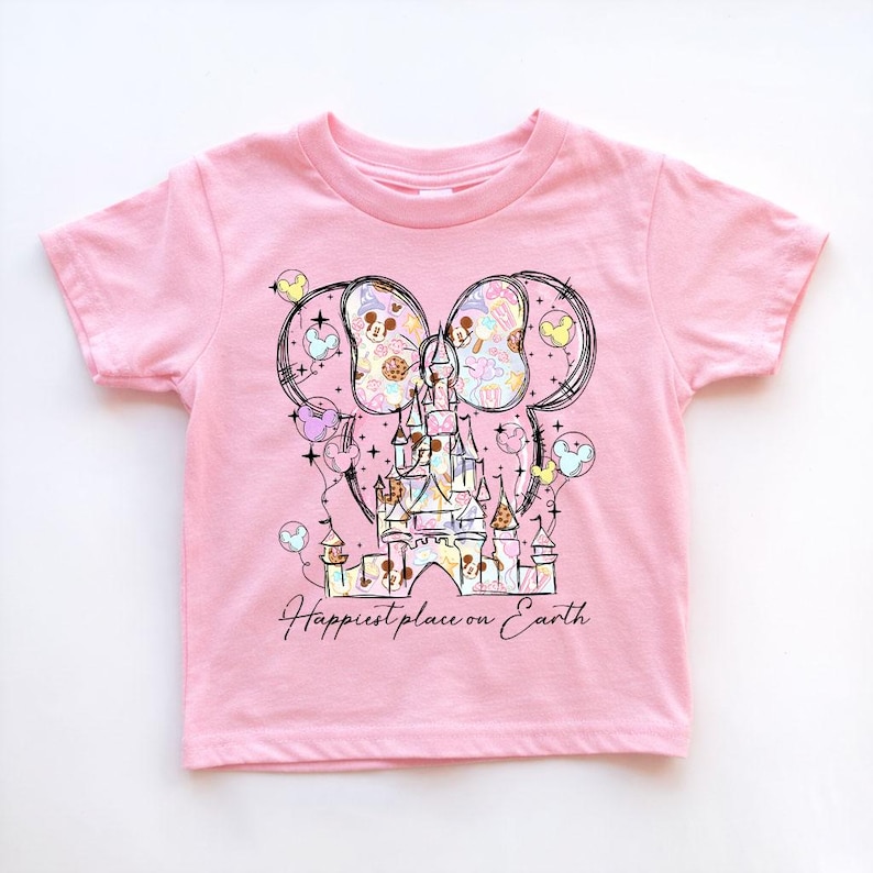 K&ouml;nnte beinhalten: Rosafarbenes T-Shirt mit kurzen &Auml;rmeln und einer Grafik eines Schlosses, Mickey Mouse-Ohren und Luftballons. Das Design enth&auml;lt den Text "Happiest place on Earth" in Schreibschrift. Die Grafik ist in Pastellfarben gehalten.