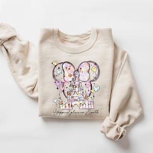 K&ouml;nnte beinhalten: Beiges Sweatshirt mit einer farbenfrohen Illustration eines Schlosses, Mickey Mouse Ohren und Luftballons. Der Text "Happiest place on Earth" steht unter der Grafik. Das Sweatshirt hat lange &Auml;rmel und einen Rundhalsausschnitt.