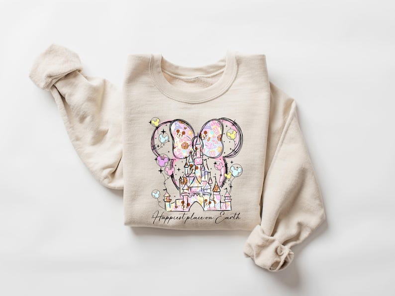 K&ouml;nnte beinhalten: Beiges Sweatshirt mit einer farbenfrohen Illustration eines Schlosses, Mickey Mouse Ohren und Luftballons. Der Text "Happiest place on Earth" steht unter der Grafik. Das Sweatshirt hat lange &Auml;rmel und einen Rundhalsausschnitt.