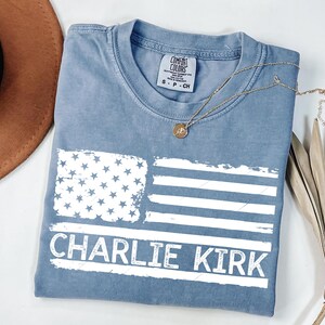 Peut inclure: Un t-shirt bleu avec un drapeau am&eacute;ricain vieilli et le nom "CHARLIE KIRK" en lettres blanches. Le t-shirt a un col rond et des manches courtes. Un collier dor&eacute; avec un pendentif est pos&eacute; sur le t-shirt.