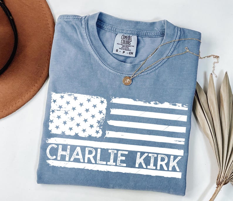 Peut inclure: Un t-shirt bleu avec un drapeau am&eacute;ricain vieilli et le nom "CHARLIE KIRK" en lettres blanches. Le t-shirt a un col rond et des manches courtes. Un collier dor&eacute; avec un pendentif est pos&eacute; sur le t-shirt.