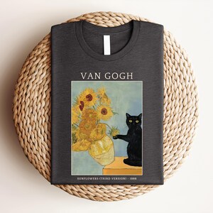 May include: Dark gray t-shirt featuring a print of Van Gogh's "Sunflowers" with a black cat. The text "VAN GOGH" is above the image, and "SUNFLOWERS (THIRD VERSION) - 1888" is below. The shirt is folded on a woven mat.