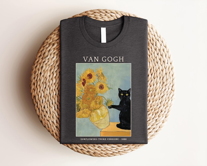 May include: Dark gray t-shirt featuring a print of Van Gogh's "Sunflowers" with a black cat. The text "VAN GOGH" is above the image, and "SUNFLOWERS (THIRD VERSION) - 1888" is below. The shirt is folded on a woven mat.