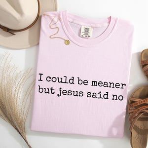 Pu&ograve; includere: Una t-shirt rosa chiaro con il testo stampato in nero I could be meaner but jesus said no. Il capo presenta uno scollo rotondo e un'etichetta del marchio Comfort Colors sul colletto interno. &Egrave; mostrata insieme a un cappello beige e sandali marroni.