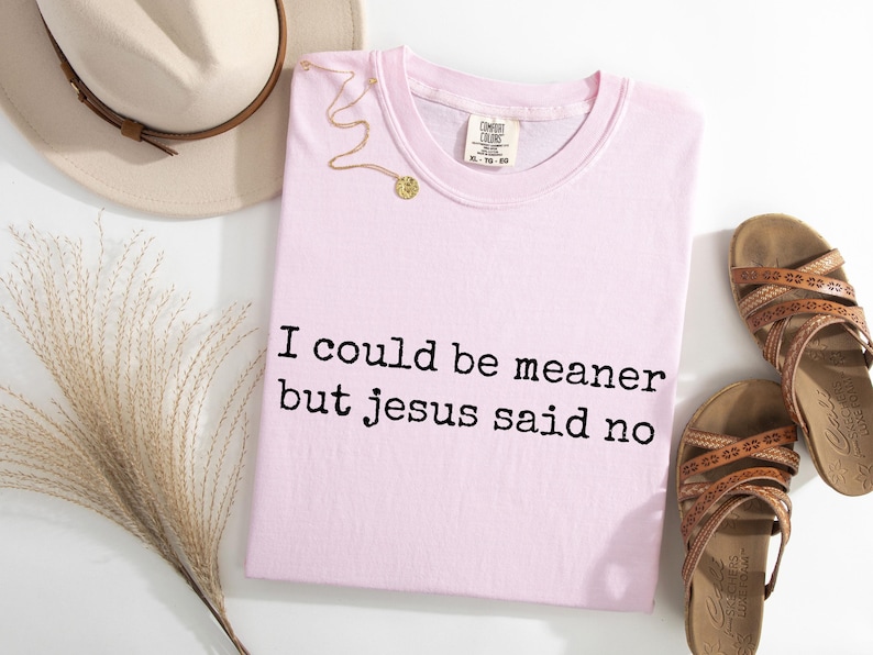 Pu&ograve; includere: Una t-shirt rosa chiaro con il testo stampato in nero I could be meaner but jesus said no. Il capo presenta uno scollo rotondo e un'etichetta del marchio Comfort Colors sul colletto interno. &Egrave; mostrata insieme a un cappello beige e sandali marroni.