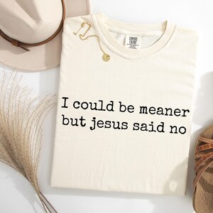 Pu&ograve; includere: Una t-shirt a maniche corte color crema con il testo nero "I could be meaner but jesus said no" stampato sul davanti. La maglietta &egrave; mostrata insieme a un cappello beige, una collana color oro e un paio di sandali marroni con cinturini.