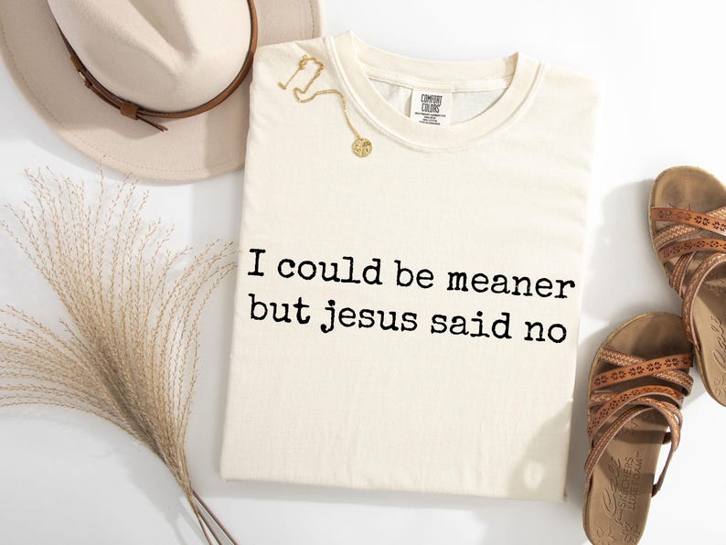 Pu&ograve; includere: Una t-shirt a maniche corte color crema con il testo nero "I could be meaner but jesus said no" stampato sul davanti. La maglietta &egrave; mostrata insieme a un cappello beige, una collana color oro e un paio di sandali marroni con cinturini.