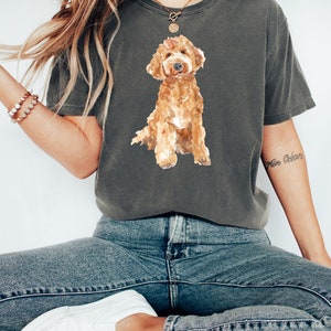 May include: A woman wearing a dark gray t-shirt with a watercolor illustration of a brown and white doodle dog. The shirt has a crew neck and short sleeves. The woman is wearing blue jeans and white sneakers.