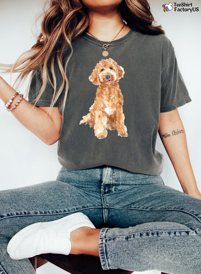 May include: A woman wearing a dark gray t-shirt with a watercolor illustration of a brown and white doodle dog. The shirt has a crew neck and short sleeves. The woman is wearing blue jeans and white sneakers.