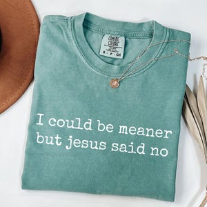 Pu&ograve; includere: Una t-shirt a maniche corte color verde salvia con scollo rotondo e la scritta bianca "I could be meaner but jesus said no" stampata sul davanti. La maglietta presenta un'etichetta del marchio Comfort Colors all'interno del collo ed &egrave; taglia S.