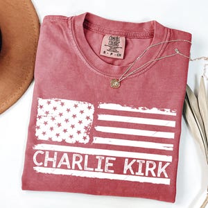 Peut inclure: Un t-shirt rouge avec un motif de drapeau am&eacute;ricain vieilli et le texte "CHARLIE KIRK" en blanc. Le t-shirt a un col rond et une petite &eacute;tiquette au niveau de l'encolure. Un collier en or avec un pendentif est drap&eacute; sur le t-shirt.
