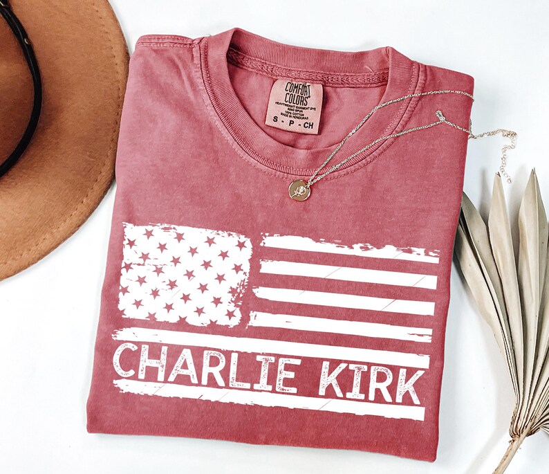 Peut inclure: Un t-shirt rouge avec un motif de drapeau am&eacute;ricain vieilli et le texte "CHARLIE KIRK" en blanc. Le t-shirt a un col rond et une petite &eacute;tiquette au niveau de l'encolure. Un collier en or avec un pendentif est drap&eacute; sur le t-shirt.