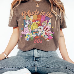 Pode incluir: Uma t-shirt castanha de manga curta com uma impress&atilde;o gr&aacute;fica com o texto "Made in the 80s" sobre um fundo arco-&iacute;ris colorido. O design inclui v&aacute;rias personagens de desenhos animados como os Care Bears, Smurfs e Muppet Babies com estrelas.