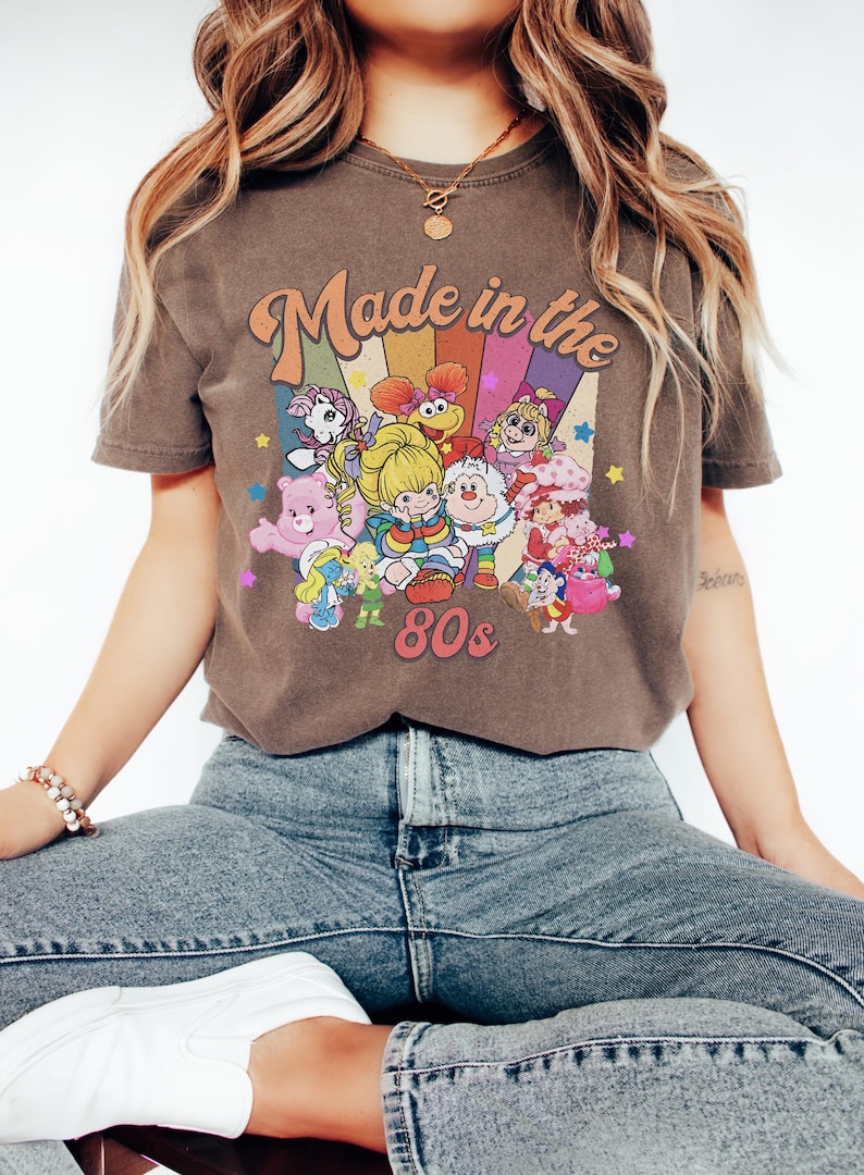 Pode incluir: Uma t-shirt castanha de manga curta com uma impress&atilde;o gr&aacute;fica com o texto "Made in the 80s" sobre um fundo arco-&iacute;ris colorido. O design inclui v&aacute;rias personagens de desenhos animados como os Care Bears, Smurfs e Muppet Babies com estrelas.
