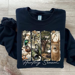 Może przedstawiać: Granatowa bluza z nadrukiem przedstawiającym kaczkę, lornetkę i jelenia na tle kamuflażu. Pod spodem białą czcionką napisano "Hunting Season".