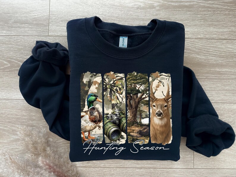 Może przedstawiać: Granatowa bluza z nadrukiem przedstawiającym kaczkę, lornetkę i jelenia na tle kamuflażu. Pod spodem białą czcionką napisano "Hunting Season".