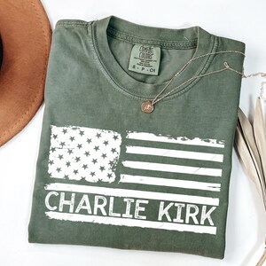 Peut inclure: T-shirt vert olive avec un drapeau am&eacute;ricain vieilli et le texte "CHARLIE KIRK" en blanc. Un collier en or avec un pendentif est pos&eacute; sur le t-shirt. Un chapeau marron et une plante s&eacute;ch&eacute;e sont en arri&egrave;re-plan.