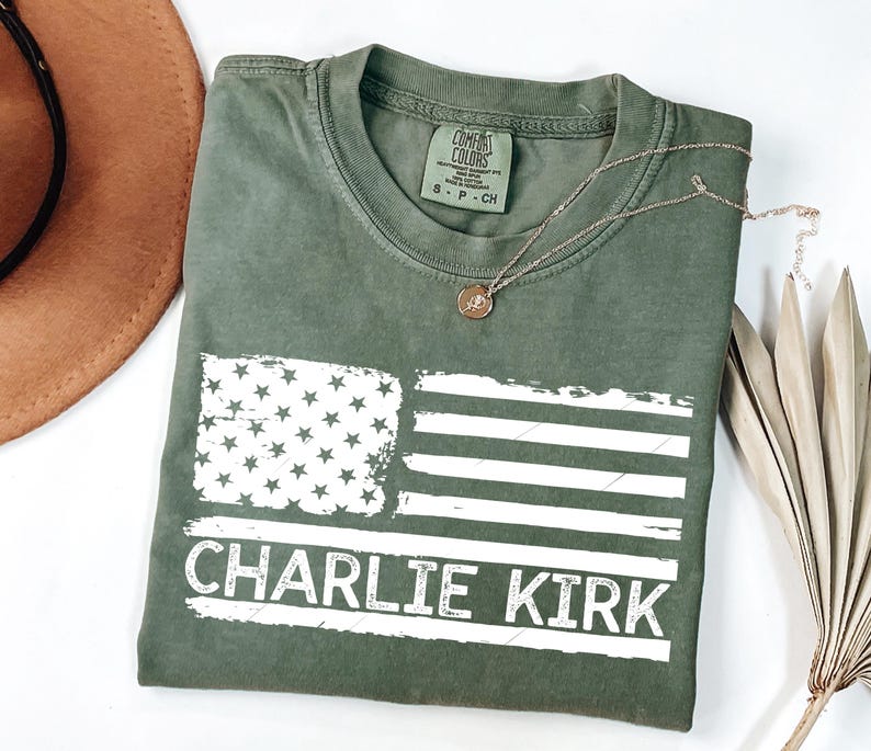 Peut inclure: T-shirt vert olive avec un drapeau am&eacute;ricain vieilli et le texte "CHARLIE KIRK" en blanc. Un collier en or avec un pendentif est pos&eacute; sur le t-shirt. Un chapeau marron et une plante s&eacute;ch&eacute;e sont en arri&egrave;re-plan.