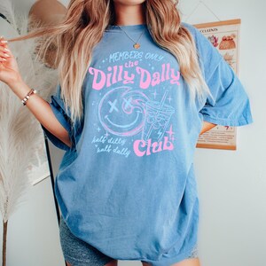 May include: A blue short-sleeve t-shirt featuring a pink and light blue graphic print. The design includes a smiley face with X-shaped eyes, a drink cup, and the text: Members Only the Dilly Dally Club, half dilly half dally.