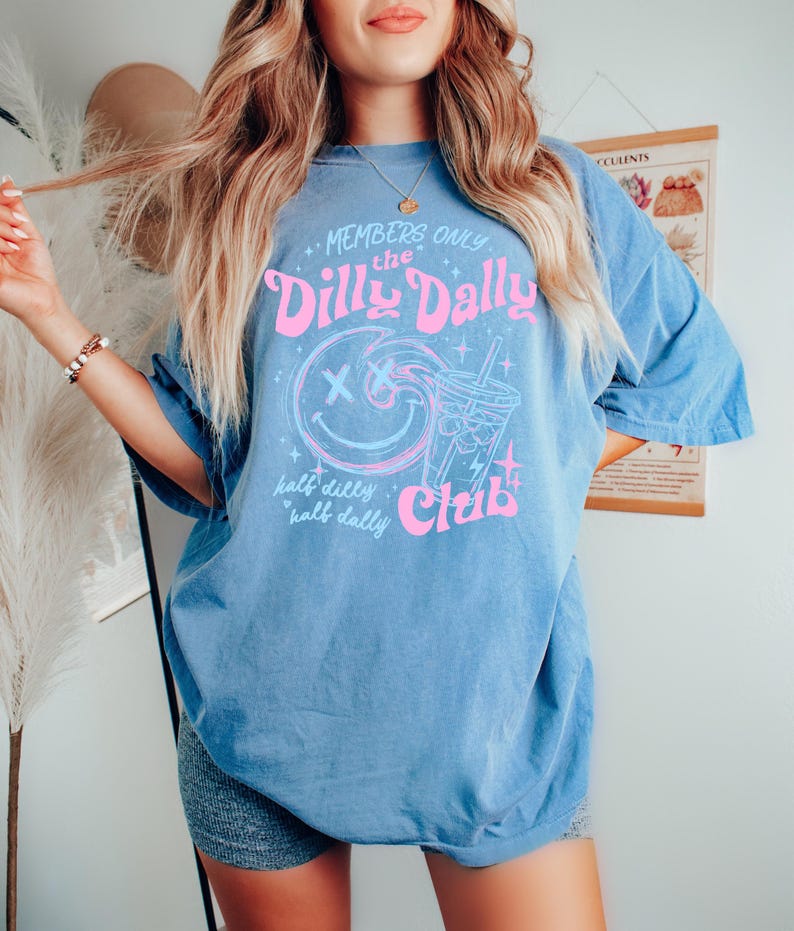 May include: A blue short-sleeve t-shirt featuring a pink and light blue graphic print. The design includes a smiley face with X-shaped eyes, a drink cup, and the text: Members Only the Dilly Dally Club, half dilly half dally.
