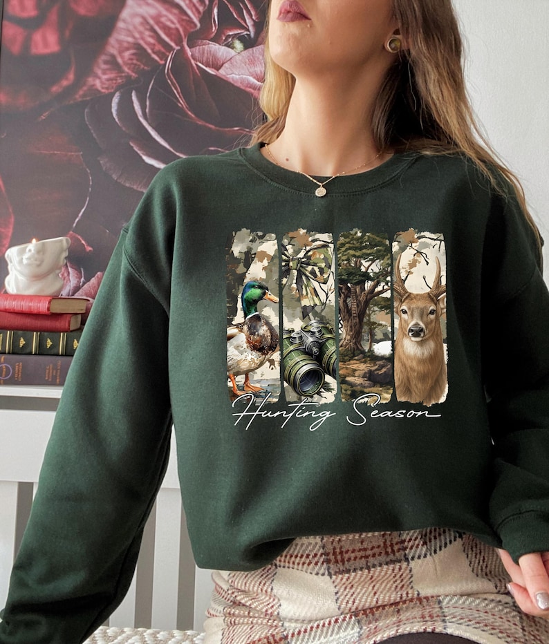 Może przedstawiać: Ciemnozielona bluza z prostokątną grafiką przedstawiającą kaczkę, lornetkę, drzewo i jelenia. Słowa "Hunting Season" są napisane białym skryptem poniżej. Bluza jest z długim rękawem.