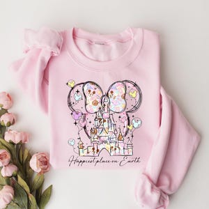K&ouml;nnte beinhalten: Hellrosa Sweatshirt mit einem skurrilen Schlossdesign mit Mickey Mouse Ohren und Luftballons. Die Grafik enth&auml;lt Pastellfarben und den Text "Happiest place on Earth". Das Sweatshirt hat einen Rundhalsausschnitt und lange &Auml;rmel.
