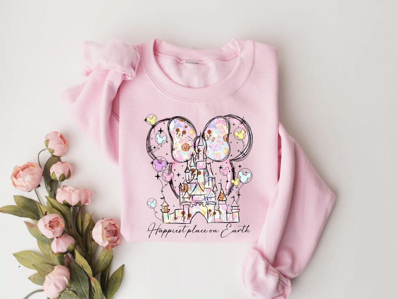 K&ouml;nnte beinhalten: Hellrosa Sweatshirt mit einem skurrilen Schlossdesign mit Mickey Mouse Ohren und Luftballons. Die Grafik enth&auml;lt Pastellfarben und den Text "Happiest place on Earth". Das Sweatshirt hat einen Rundhalsausschnitt und lange &Auml;rmel.