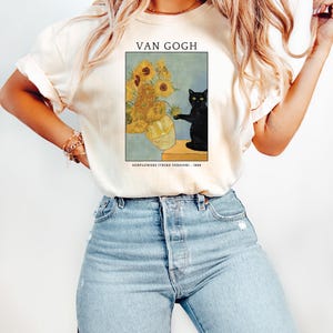 May include: Cream-colored t-shirt featuring a print of Van Gogh's "Sunflowers (Third Version)" with a black cat. The artwork includes a vase of sunflowers and the text "VAN GOGH" and "SUNFLOWERS (THIRD VERSION) - 1888".
