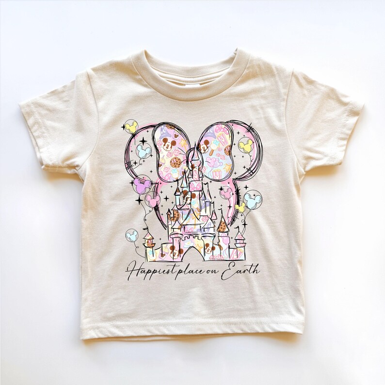 K&ouml;nnte beinhalten: Cremefarbenes Kleinkind-T-Shirt mit einer farbenfrohen Grafik eines Schlosses, Mickey Mouse-Ohren und Luftballons. Das Design enth&auml;lt den Text "Happiest place on Earth" in einer Schreibschrift. Die Grafik ist in Pastellfarben gehalten.