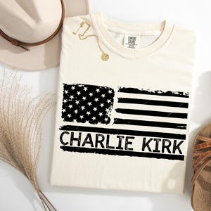 Peut inclure: T-shirt cr&egrave;me avec un motif de drapeau am&eacute;ricain noir et blanc vieilli. Le texte "CHARLIE KIRK" est imprim&eacute; en dessous. Un collier dor&eacute;, un chapeau et des sandales sont &eacute;galement visibles.