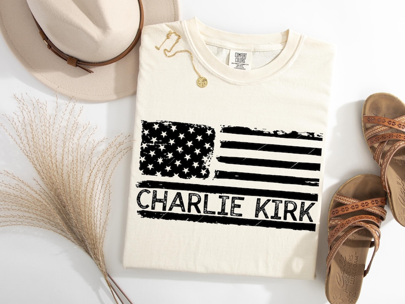 Peut inclure: T-shirt cr&egrave;me avec un motif de drapeau am&eacute;ricain noir et blanc vieilli. Le texte "CHARLIE KIRK" est imprim&eacute; en dessous. Un collier dor&eacute;, un chapeau et des sandales sont &eacute;galement visibles.