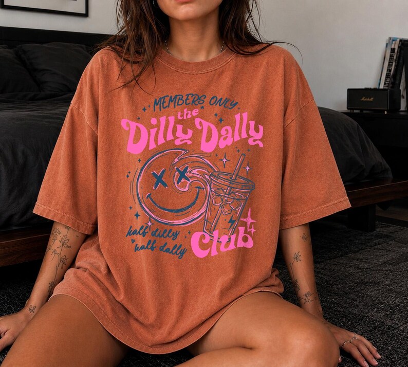 May include: A rust-colored short-sleeve t-shirt featuring a graphic print with the text "MEMBERS ONLY the Dilly Dally Club half dilly half dally". The design includes a swirling smiley face with X-shaped eyes and a drink cup with a straw.