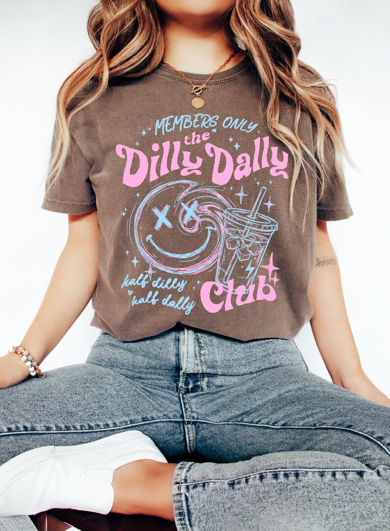 May include: A dark brown short-sleeve t-shirt featuring a graphic print with the text "MEMBERS ONLY the Dilly Dally Club half dilly half dally". The design includes a blue and pink smiley face with X eyes and a blue iced drink cup illustration.