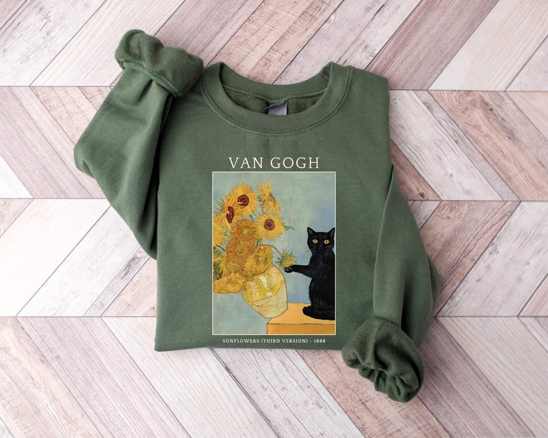 May include: Olive green sweatshirt featuring a print of Van Gogh's "Sunflowers" with a black cat. The text "VAN GOGH" is above the image, and "SUNFLOWERS (THIRD VERSION) - 1888" is below. The sweatshirt has a crew neck and long sleeves.