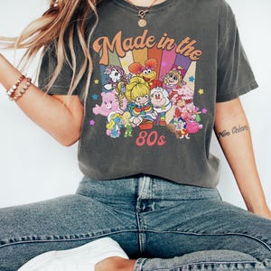 Pode incluir: Uma t-shirt de manga curta cinzento-carv&atilde;o com uma impress&atilde;o gr&aacute;fica que inclui o texto Made in the 80s. O design apresenta um fundo de arco-&iacute;ris colorido e v&aacute;rias personagens de desenhos animados, incluindo um Smurf e um Ursinho Carinhoso.
