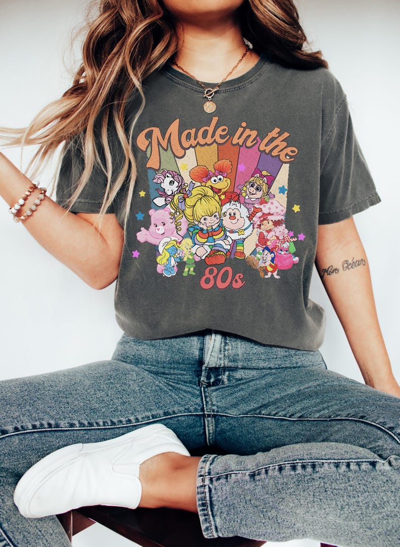 Pode incluir: Uma t-shirt de manga curta cinzento-carv&atilde;o com uma impress&atilde;o gr&aacute;fica que inclui o texto Made in the 80s. O design apresenta um fundo de arco-&iacute;ris colorido e v&aacute;rias personagens de desenhos animados, incluindo um Smurf e um Ursinho Carinhoso.