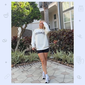 May include: A cream-colored crewneck sweatshirt featuring the word PHARMACY printed in dark navy blue block letters across the chest. The sweatshirt is paired with black athletic shorts, white crew socks, and white athletic sneakers with grey accents.