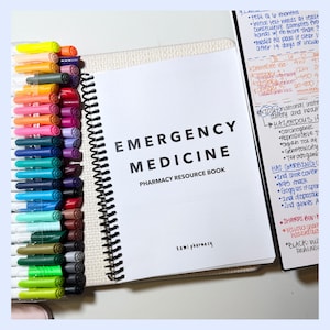 May include: A pharmacy resource book titled "Emergency Medicine" with a spiral binding. The book is accompanied by a selection of colourful markers. The text on the cover is in bold black letters. The markers are in various shades.
