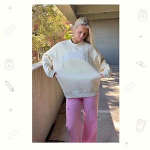 May include: A cream-colored long-sleeve sweatshirt featuring the word PHARMACY in a large, arched, white embroidered font across the chest, paired with solid light pink scrub pants with a visible side pocket and a drawstring waist.